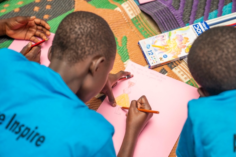 Young participants in blue shirts drawing on pink paper with pencils.