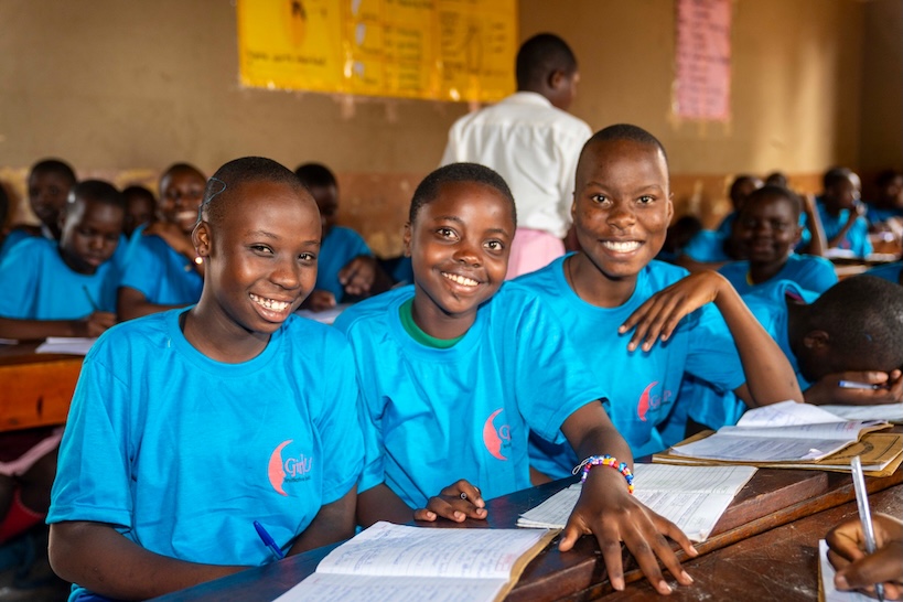 Three smiling students in blue Girl Up Initiative Uganda shirts seated at desks with exercise books in a classroom.