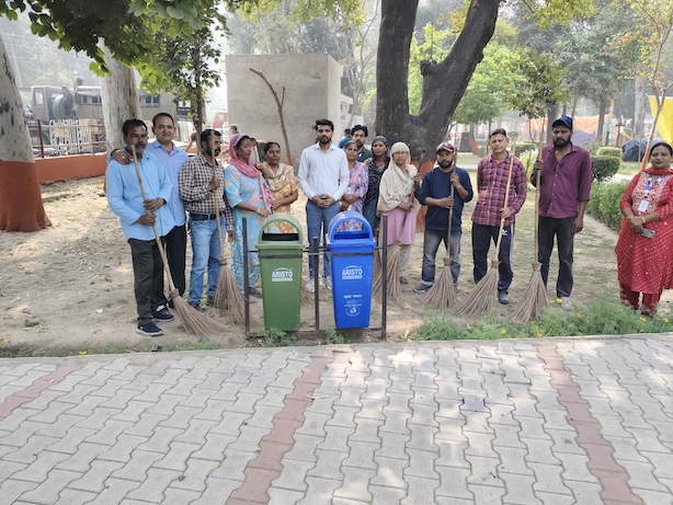 A group stands outdoors beside green and blue waste bins, with cleaning tools.