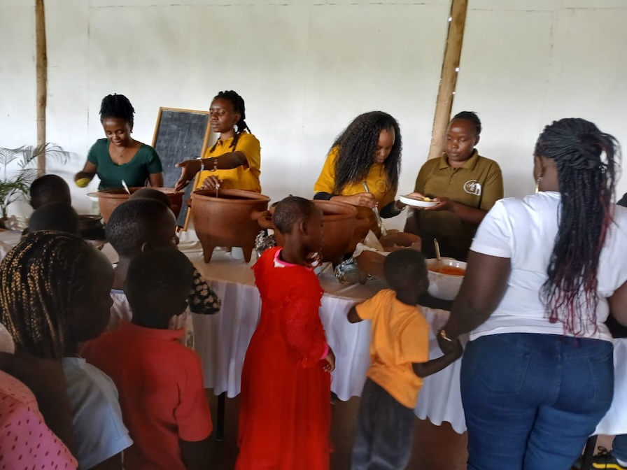 Adults serving food to children gathered around a table.