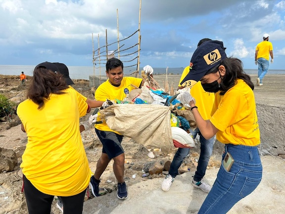 Volunteers in yellow shirts gather discarded plastic into a large sack on a beach.