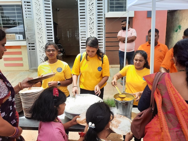 Three volunteers serve food from large containers to people gathered around a table.