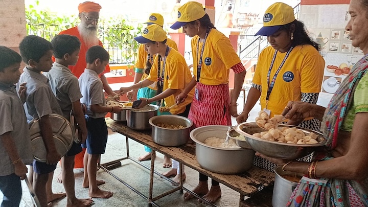 Volunteers in yellow shirts serve food to children holding plates.