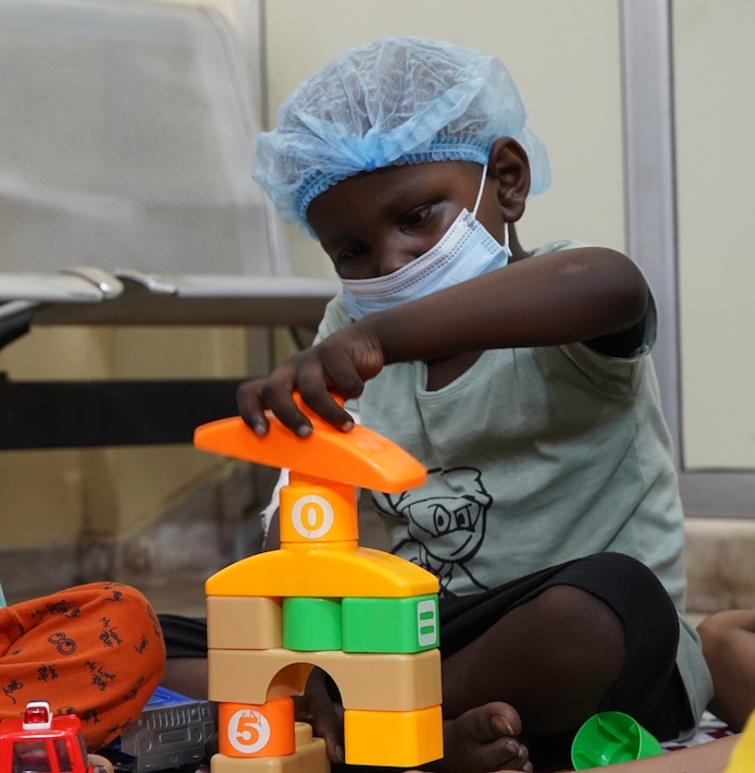 A seated child wearing a mask and protective cap stacks colourful blocks.