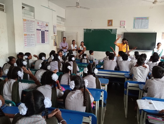 Students seated at classroom desks face a facilitator beside a blackboard.